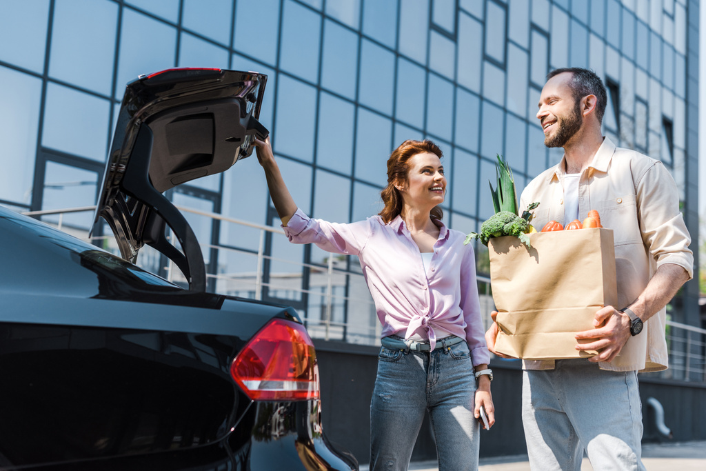 низкий угол зрения счастливой женщины, смотрящей на мужчину с бумажным пакетом, стоящим рядом с машиной
  - Фото, изображение
