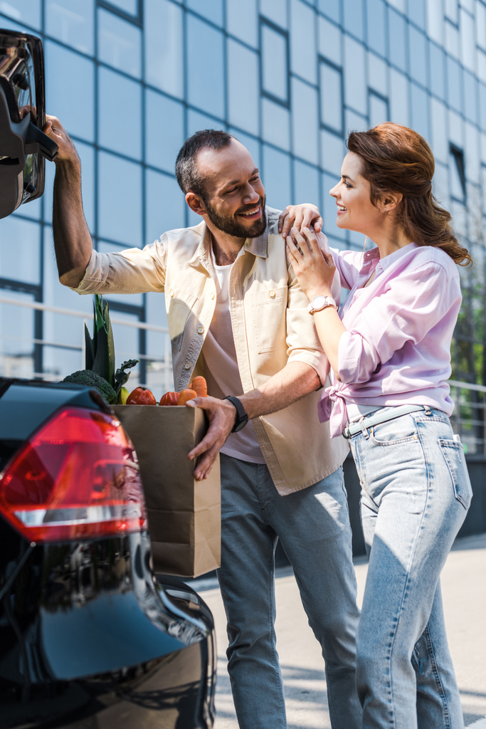 happy bearded man putting paper bag in car trunk near attractive woman  - Photo, Image