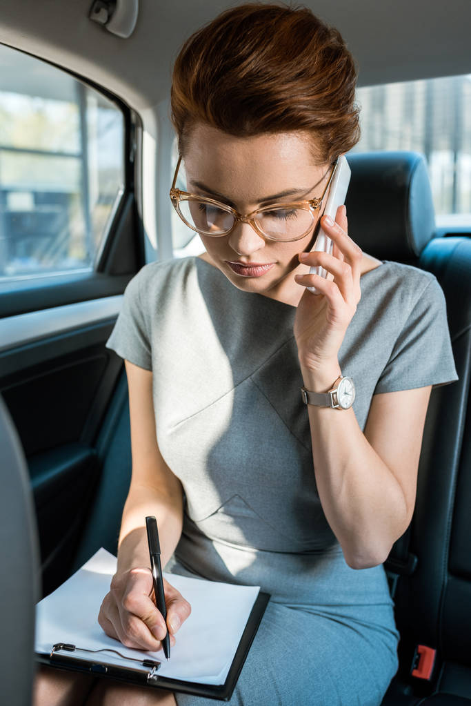 ελκυστική επιχειρηματίας σε γυαλιά γράφοντας ενώ μιλάτε στο smartphone στο αυτοκίνητο  - Φωτογραφία, εικόνα