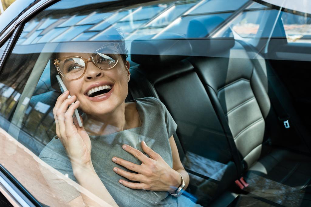 счастливая женщина в очках смеется, разговаривая по смартфону в машине
  - Фото, изображение