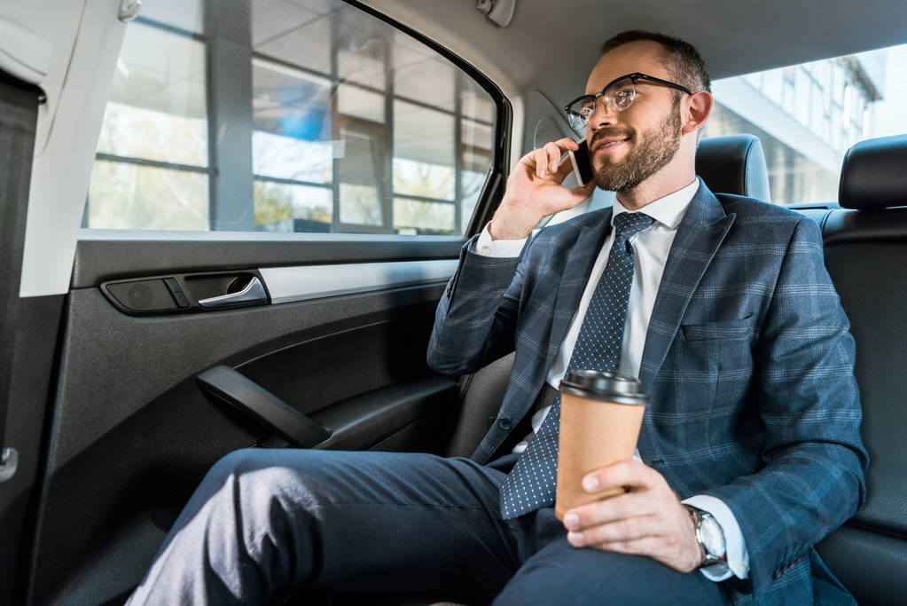 スマートフォンで話し、車の中で紙コップを持っている幸せなビジネスマンの低角度ビュー  - 写真・画像