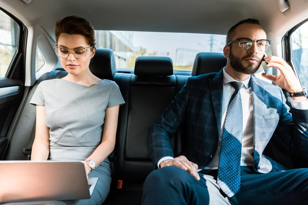 ελκυστική επιχειρηματίας χρησιμοποιώντας φορητό υπολογιστή κοντά επιχειρηματίας μιλάει στο smartphone στο αυτοκίνητο  - Φωτογραφία, εικόνα