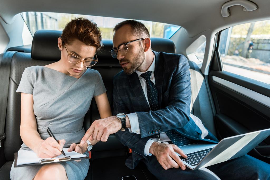 przystojny biznesmen wskazując palcem w schowku, siedząc z laptopem w pobliżu kobiety w samochodzie  - Zdjęcie, obraz
