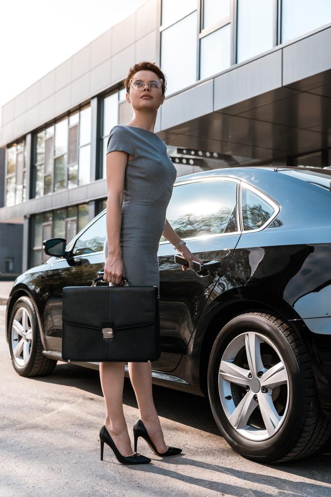 ブリーフケース付きの黒い車の近くに立っている魅力的なビジネスウーマンの低角度図  - 写真・画像