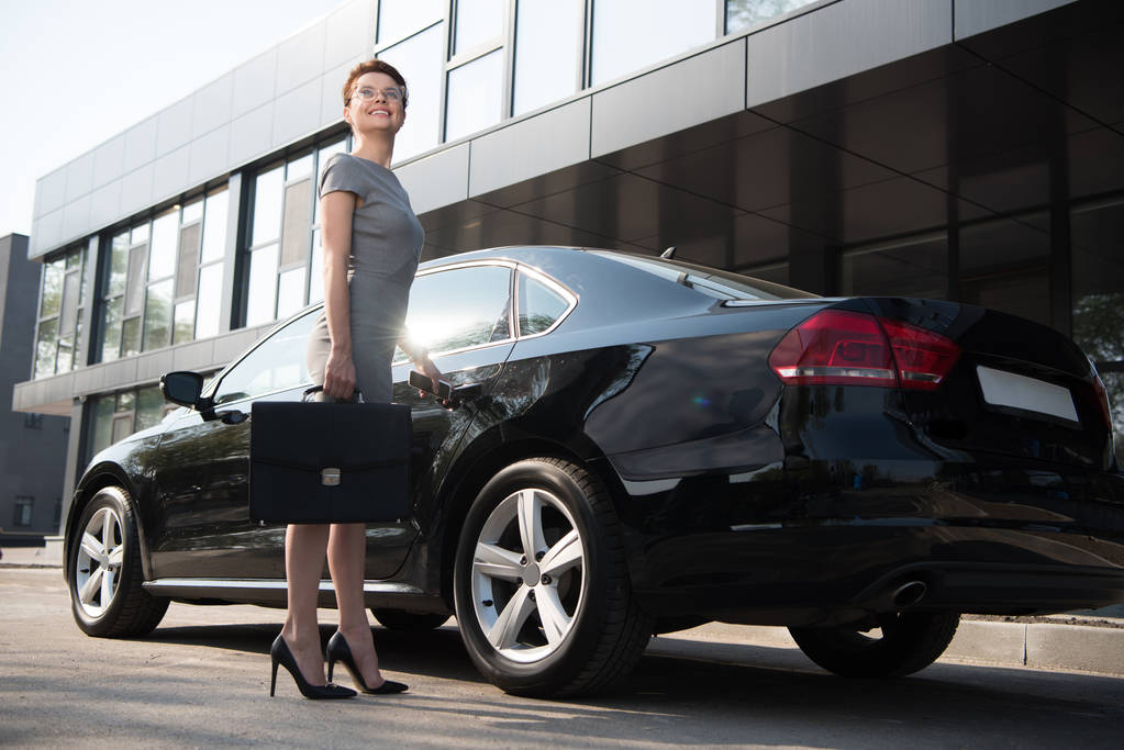 lage hoek weergave van gelukkige zakenvrouw staande in de buurt van zwarte auto met aktetas  - Foto, afbeelding