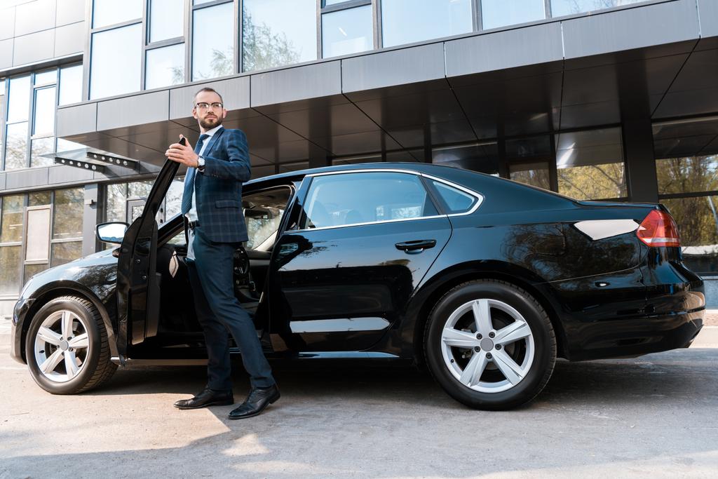 низький кут зору красивого бізнесмена в костюмі, що стоїть біля чорного автомобіля
  - Фото, зображення