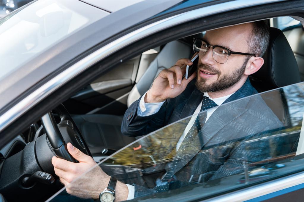 pohledný obchodník s brýlemi přidržením volantu a mluvení na smartphone  - Fotografie, Obrázek