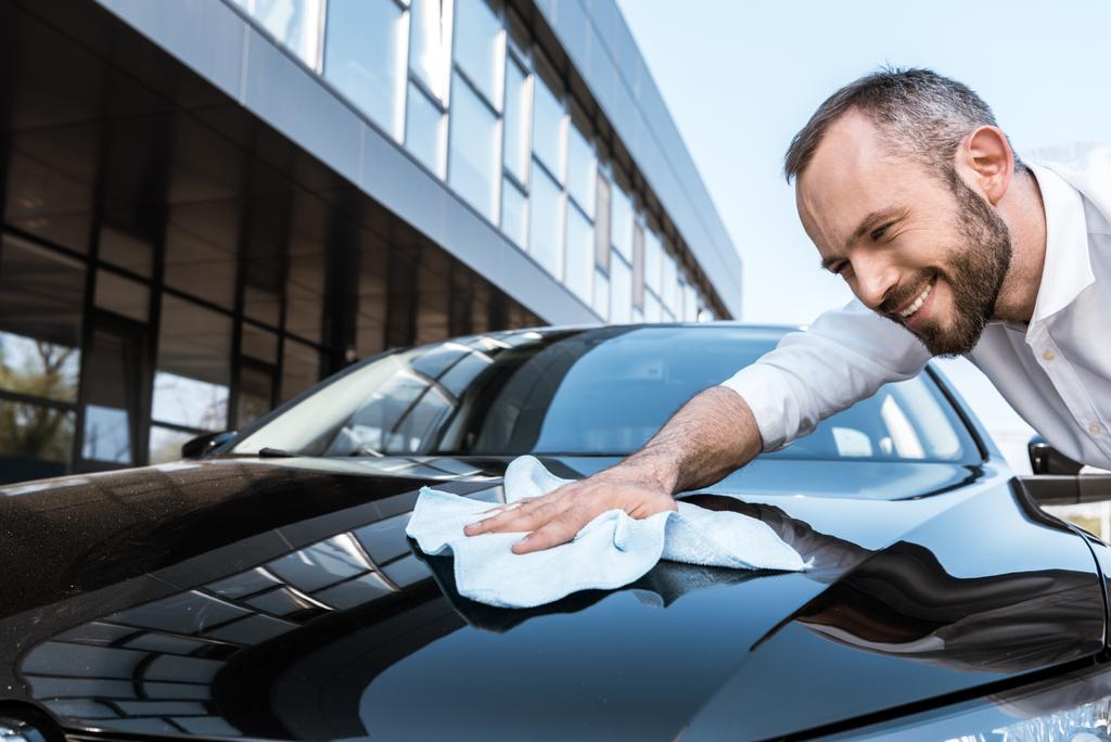 建物の近くに白い布で黒い車を掃除する幸せなビジネスマンの低角度図  - 写真・画像
