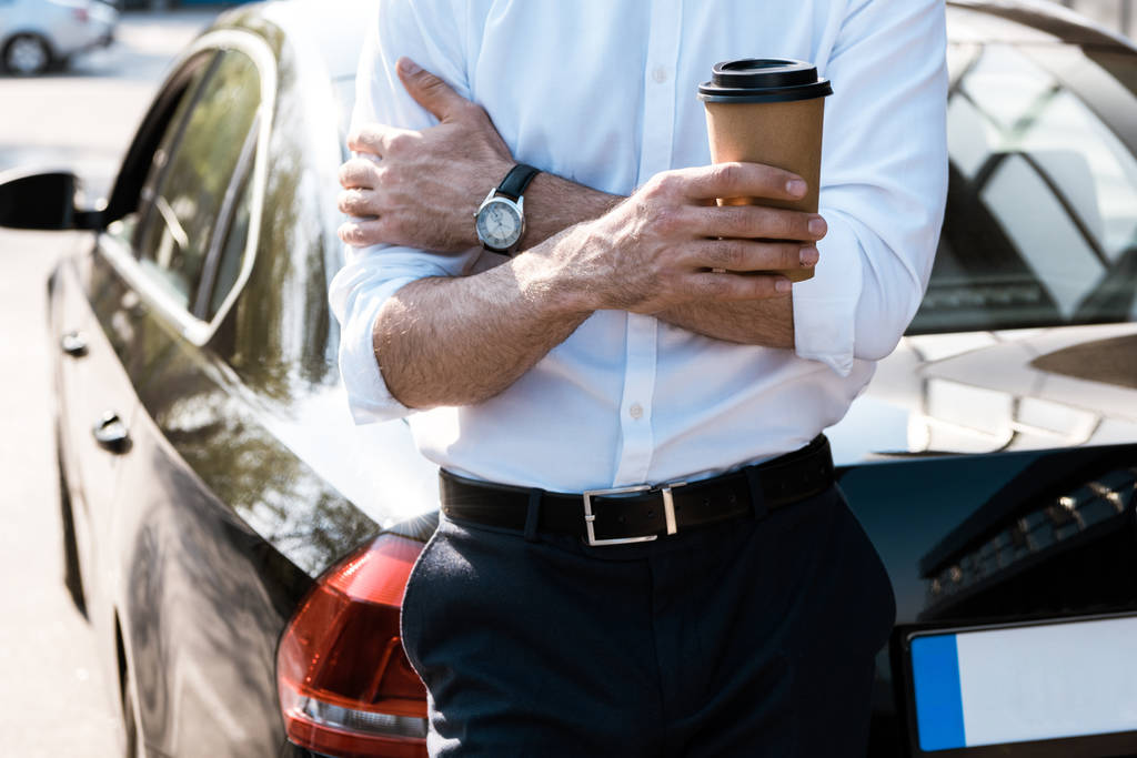 περικομμένη όψη του ανθρώπου που κρατά το χάρτινο κύπελλο ενώ στέκεται ενώ σταυρωτά χέρια κοντά στο αυτοκίνητο  - Φωτογραφία, εικόνα