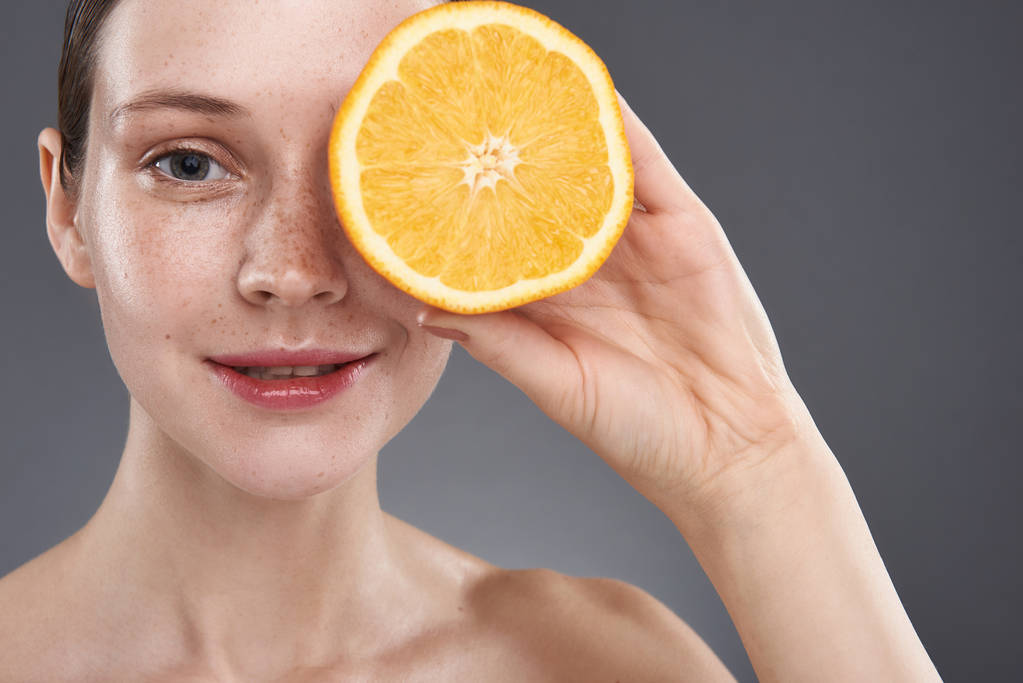 Beautiful young woman with freckles covering eye with orange - Photo, Image