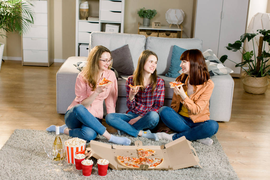 Freundschaft, Urlaub, Fast Food und Festkonzept - fröhliche junge Freundinnen mit Getränken und Popcorn, die zu Hause Pizza essen, auf dem Fußboden im gemütlichen Zimmer sitzen - Foto, Bild