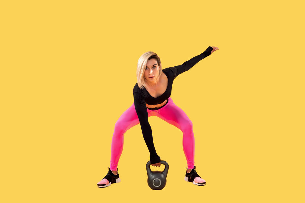 Fitness vrouw in modieuze roze en zwarte sportkleding uit te werken met kettlebell op gele achtergrond. - Foto, afbeelding