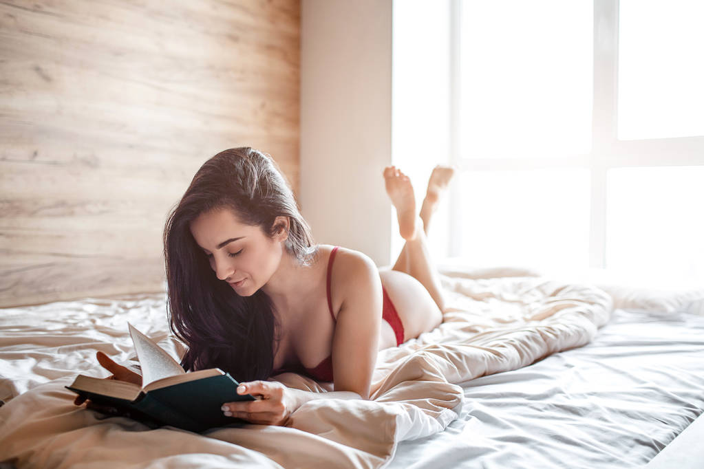 朝ベッドの上で魅惑的な若い裸の黒髪の女性。ホットセクシーなモデル嘘と読書本。穏やかで平和だ陽気なよく構築されたモデル。魅力的な人. - 写真・画像