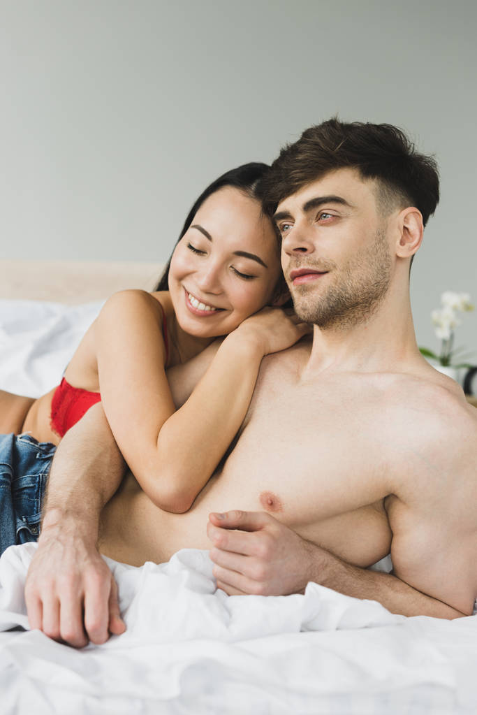 happy asian woman embracing pensive shirtless boyfriend in bedroom - Photo, Image