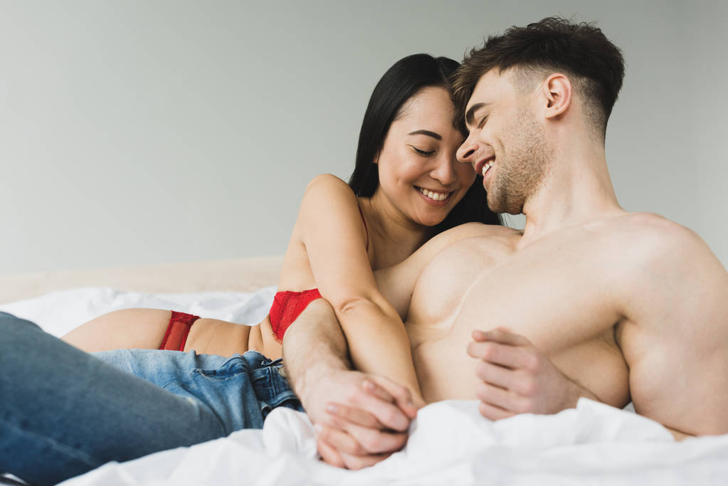 joyeux couple interracial souriant et tenant la main tout en étant couché sur le lit
 - Photo, image