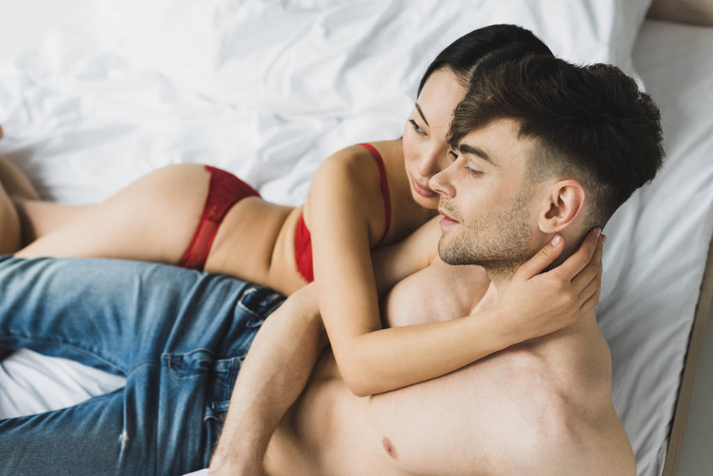 mladé mezirasové páry objímající a dívající se pryč, zatímco leží na posteli - Fotografie, Obrázek
