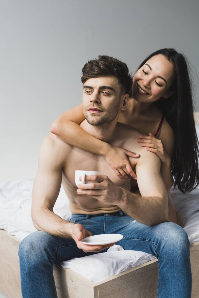 smiling asian woman hugging pensive boyfriend sitting on bed with cup of coffee - Photo, Image