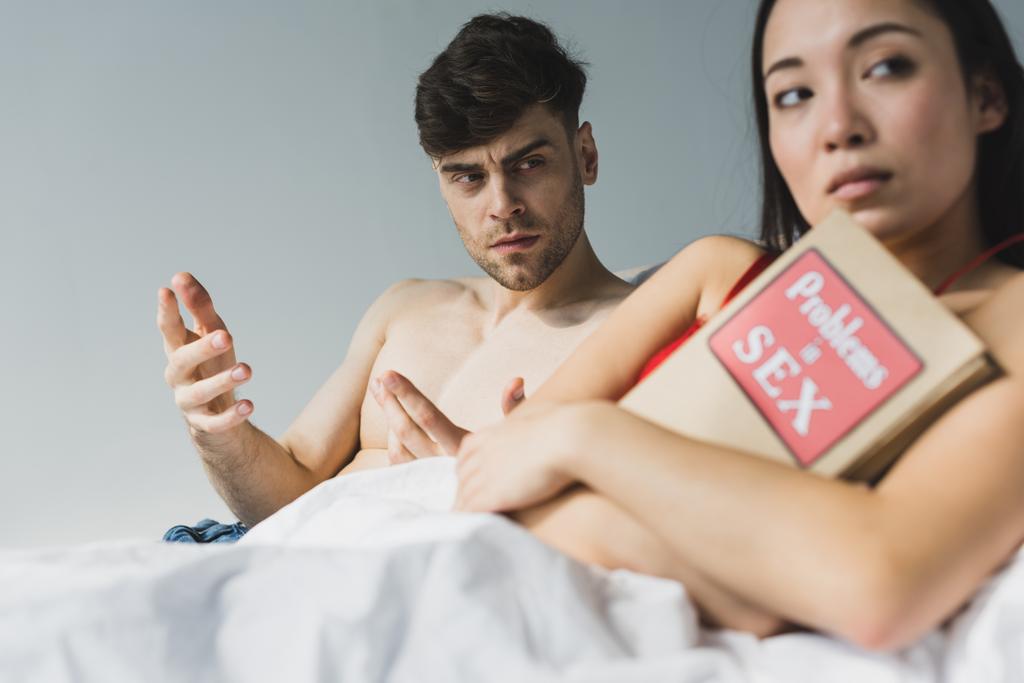 selective focus of offended asian woman holding problems in sex book while lying near dissatisfied boyfriend - Photo, Image