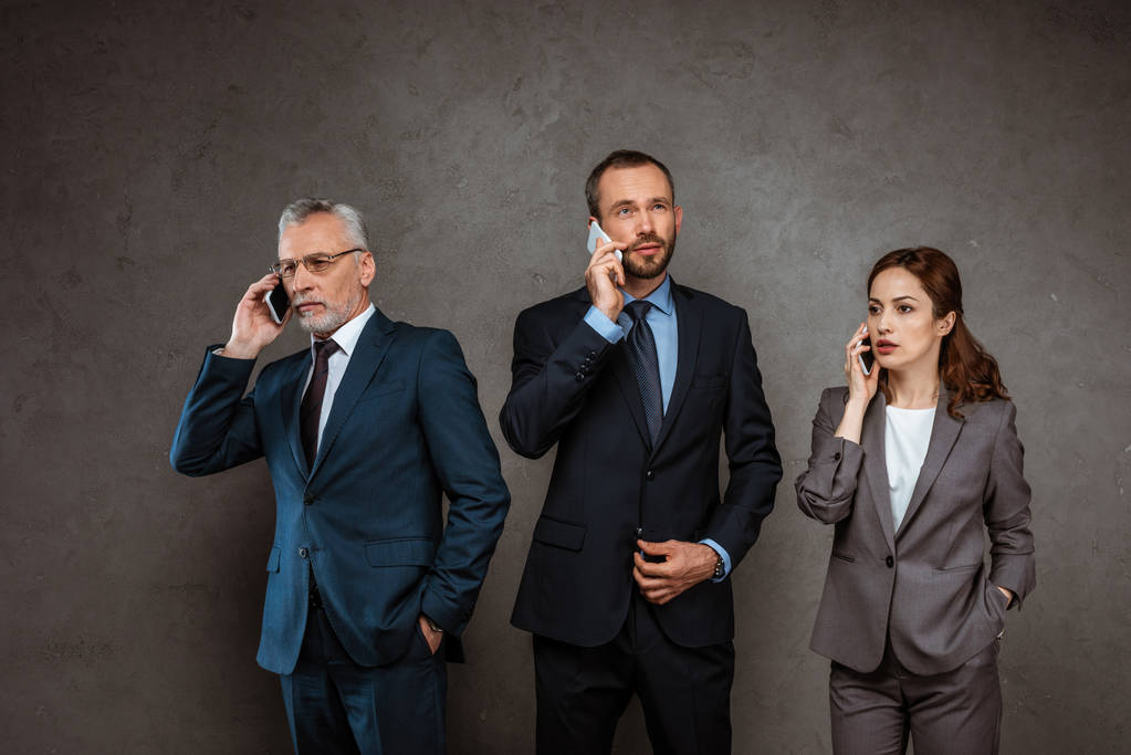 красиві бізнесмени і приваблива бізнес-леді в формальному одязі розмовляють на смартфонах на сірому
  - Фото, зображення