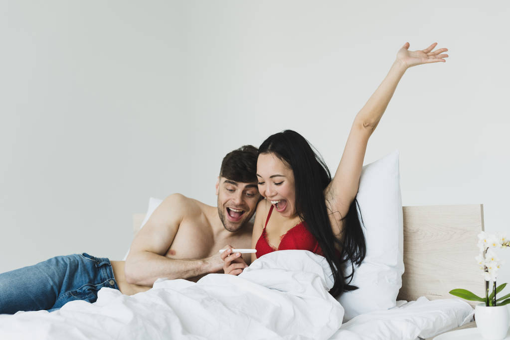 happy asian woman gesturing while holding pregnancy test near smiling boyfriend  - Photo, Image