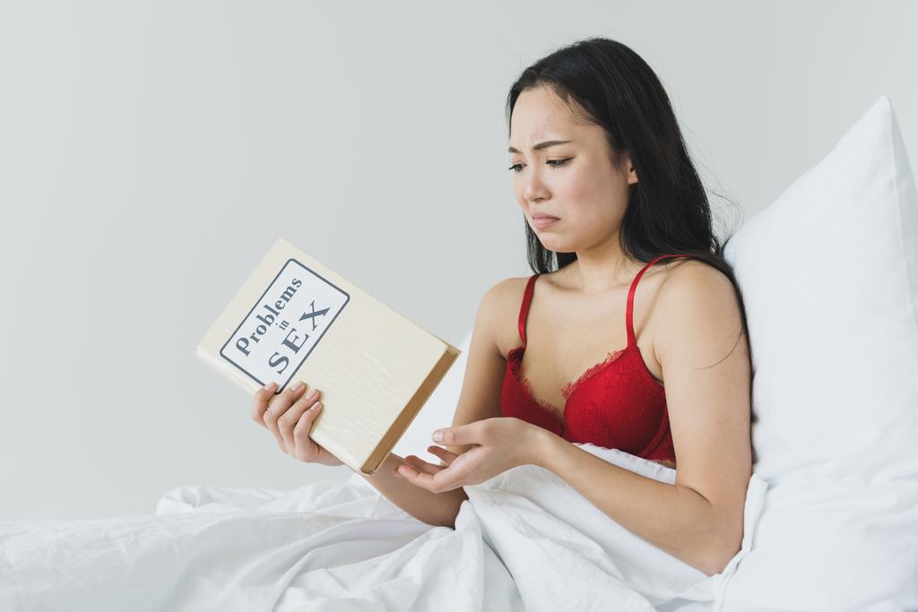 upset asian woman holding problems in sex book while sitting under white blanket  - Photo, Image