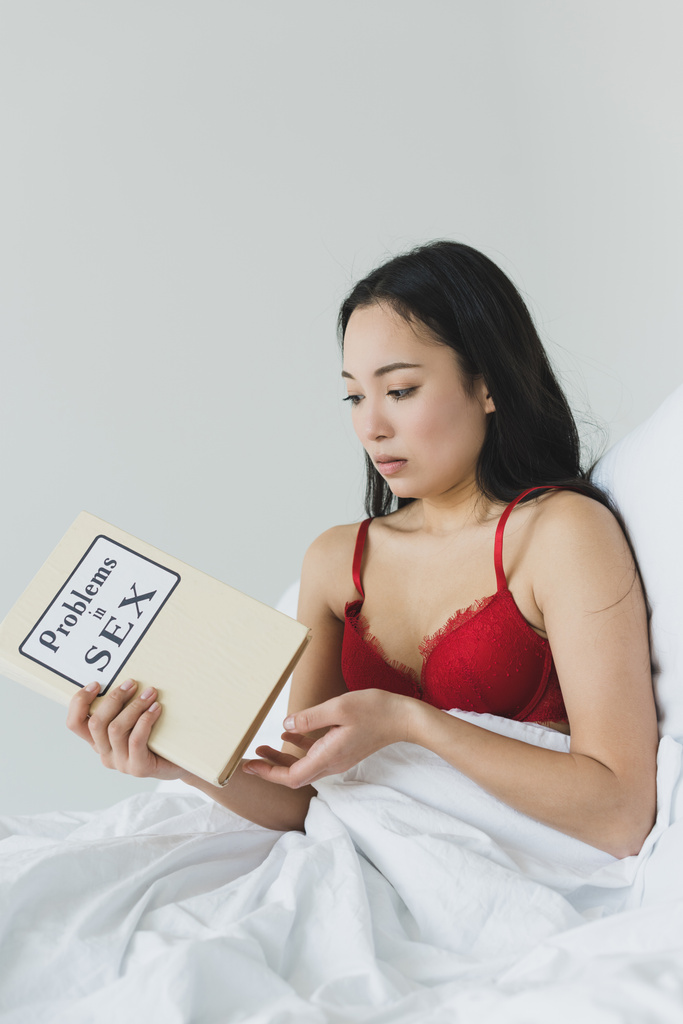 удивлена азиатская женщина, держащая проблемы в книге секса, пока лежит в постели
 - Фото, изображение