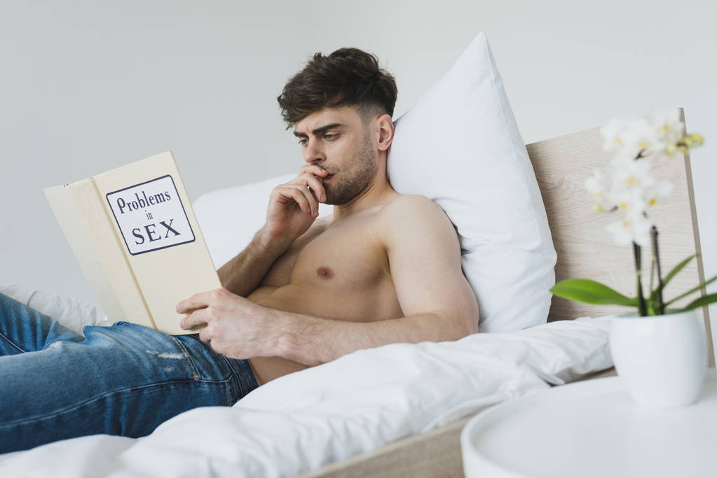 pensativo bonito homem leitura problemas no sexo livro enquanto deitado na cama no azul jeans
 - Foto, Imagem
