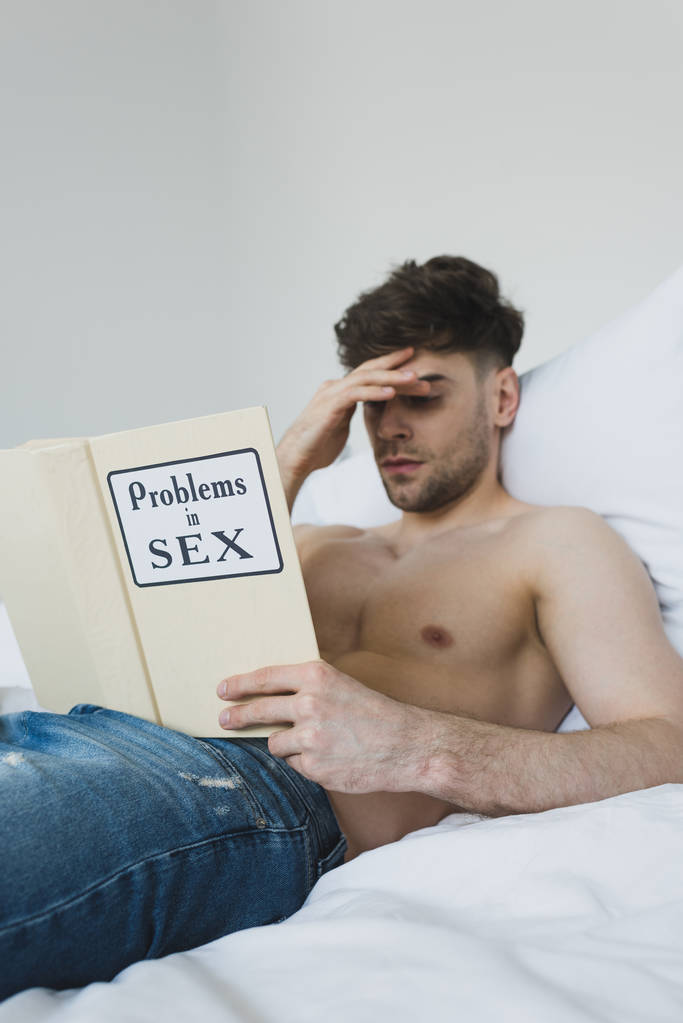 serious shirtless man reading problems in sex book while lying in bed in blue jeans - Photo, Image