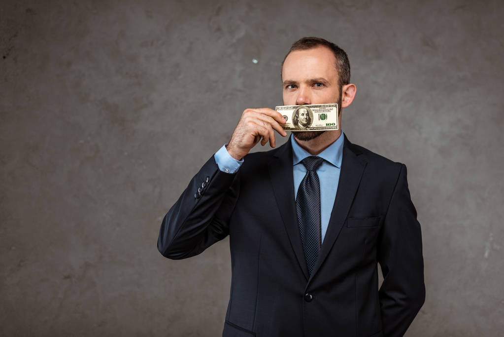 Geschäftsmann im Anzug bedeckt Mund mit Dollarschein, während er auf grau steht  - Foto, Bild