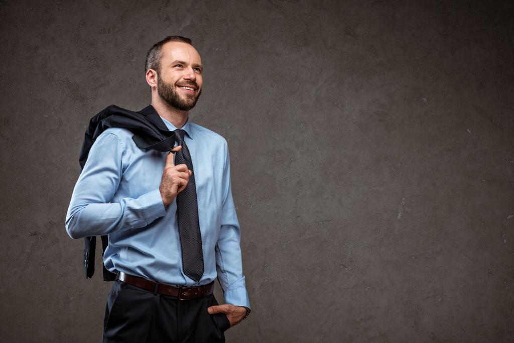 χαρούμενος επιχειρηματίας στέκεται με το χέρι στην τσέπη και χαμογελά σε γκρι  - Φωτογραφία, εικόνα