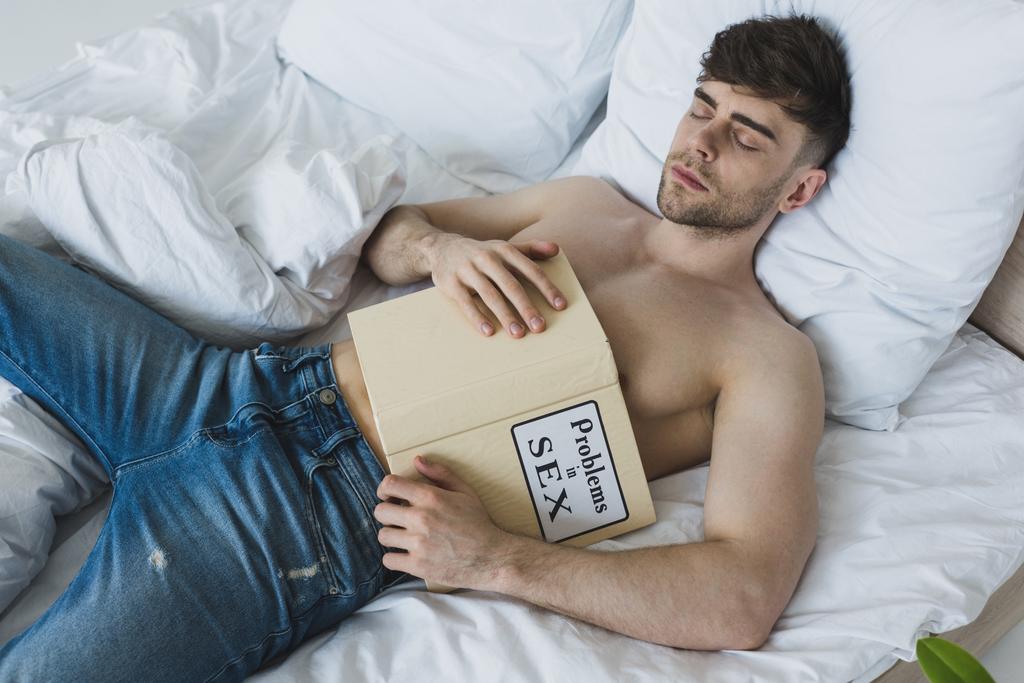knap shirtless man het houden van problemen in seks boek terwijl liggend op witte beddengoed in blauwe jeans - Foto, afbeelding