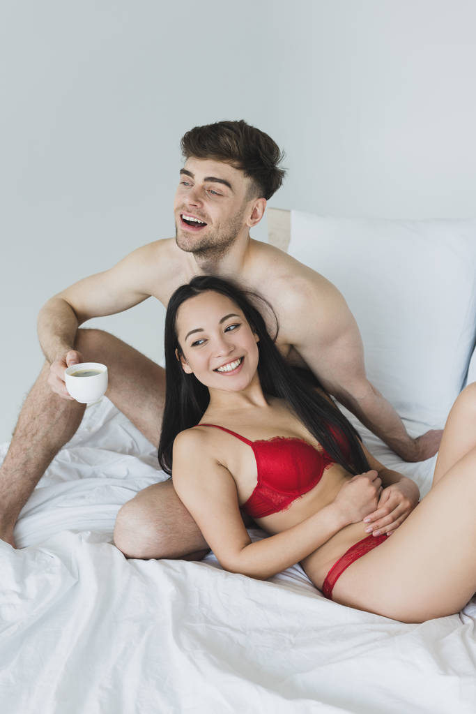 uśmiechnięty mężczyzna trzymając filiżankę kawy siedząc na białym łóżku w pobliżu wesoły Asian Girlfriend - Zdjęcie, obraz