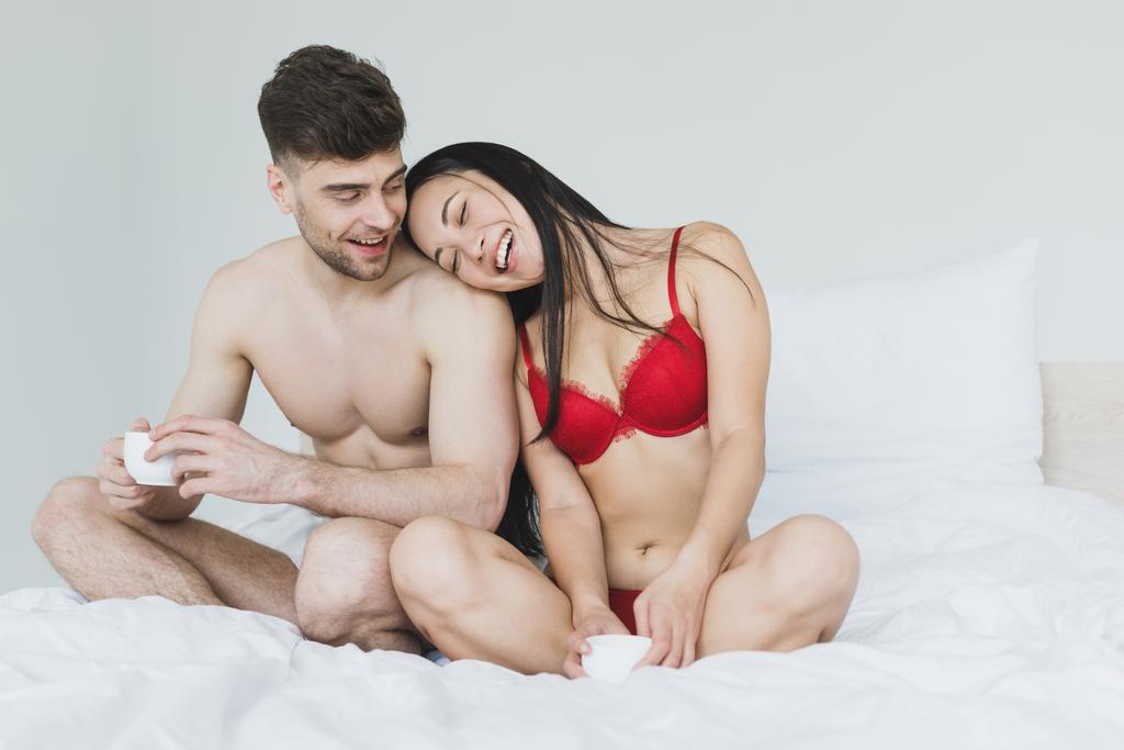 szczęśliwy Interracial para siedzi na łóżku z skrzyżowanymi nogami i trzymając filiżanki kawy  - Zdjęcie, obraz