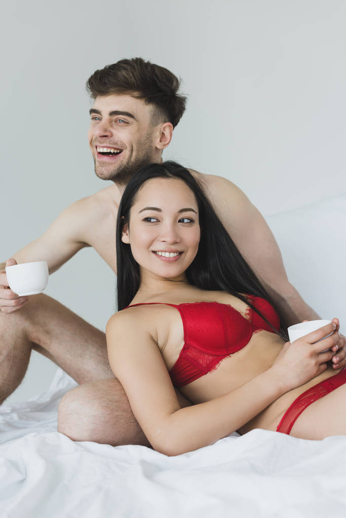 vrolijke shirtless man met koffie kopje terwijl zittend op wit beddengoed in de buurt van sexy lachende Aziatische vriendin - Foto, afbeelding