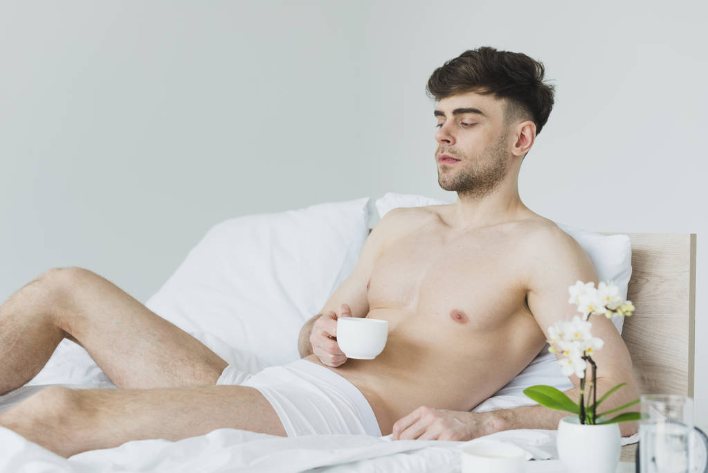 γοητευτικός όμορφος άνθρωπος κρατώντας τον άνθρωπο κρατώντας κούπα καφέ, ενώ ξαπλωμένοι σε λευκό κρεβάτι με εσώρουχα - Φωτογραφία, εικόνα