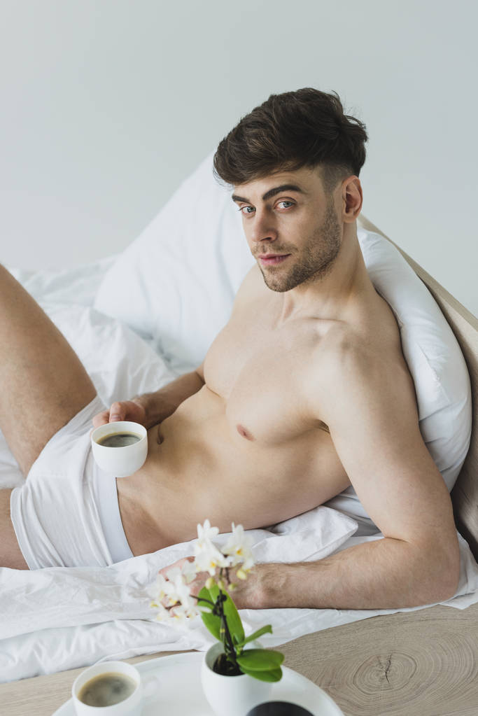 下着姿の白い寝具の上に横たわって、コーヒーカップを持ち、カメラを見ているハンサムな男 - 写真・画像