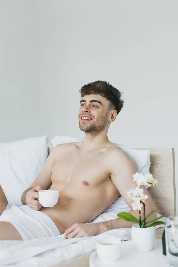 veselý muž ve spodním prádle s šálkem kávy, zatímco leží v posteli a dívá se jinam - Fotografie, Obrázek