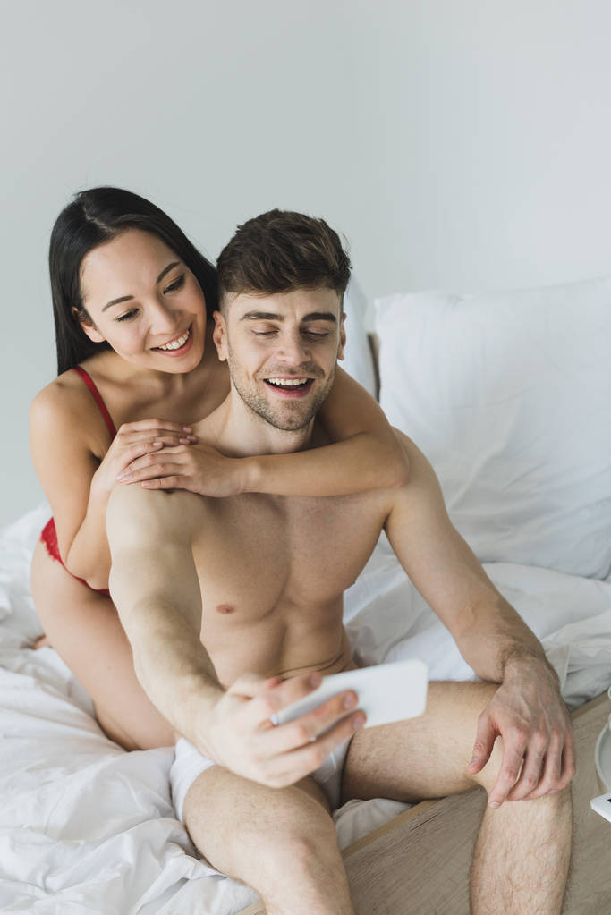 白い寝具の上に座って、スマートフォンで自分撮りを取る下着で幸せな異人種間のカップル - 写真・画像