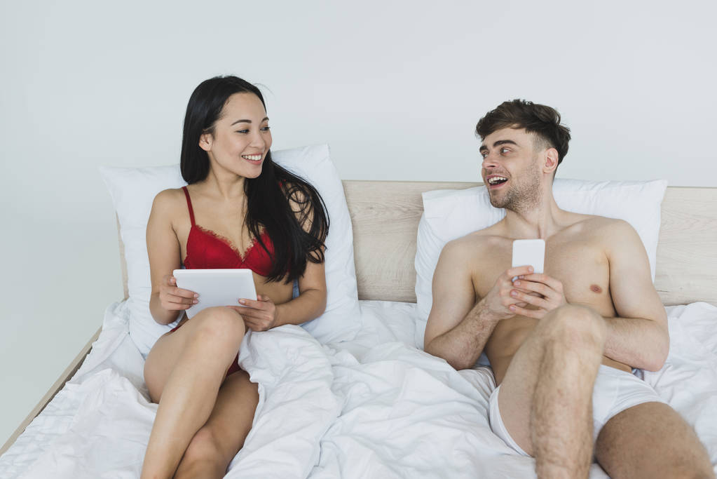 sorrindo casal interracial usando tablet digital e smartphone enquanto deitado na cama e olhando um para o outro
 - Foto, Imagem