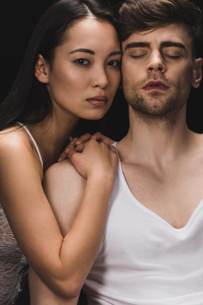 aantrekkelijke Aziatische vrouw liggend op schouder van knappe vriendje en kijken naar camera geïsoleerd op zwart - Foto, afbeelding