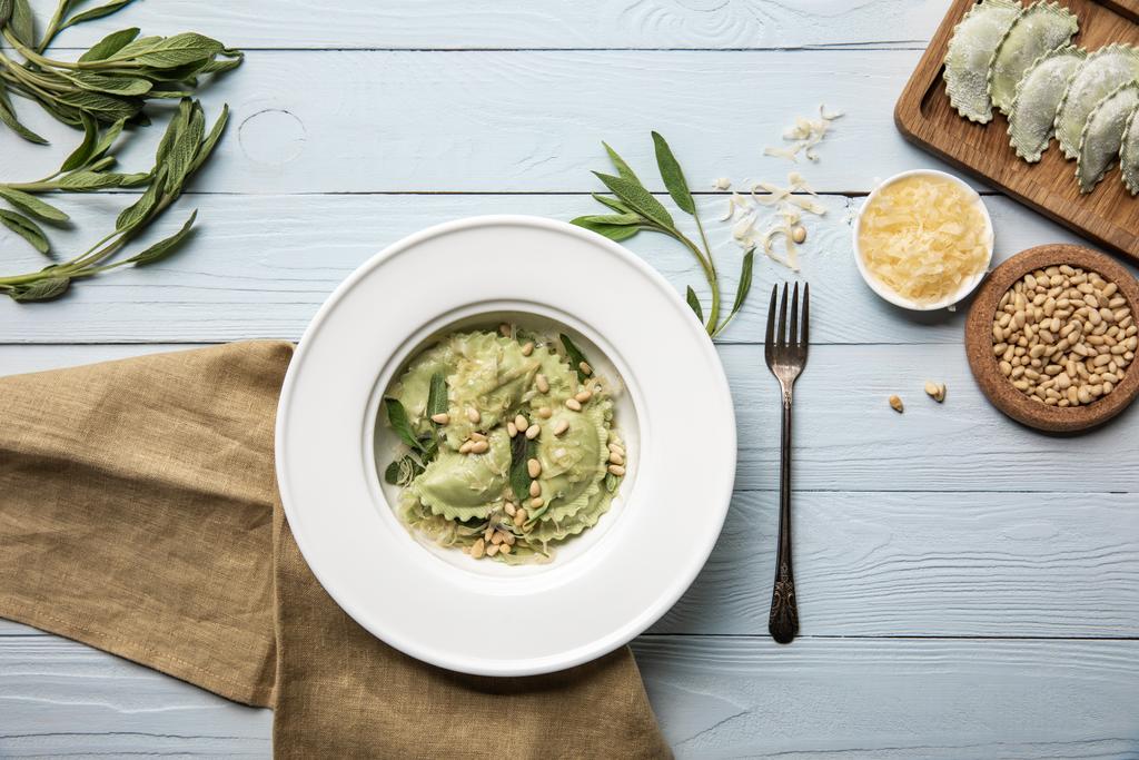 top view of green ravioli served in white plate with pine nuts, sage and grated cheese on white wooden table with fork and sackcloth - Photo, Image
