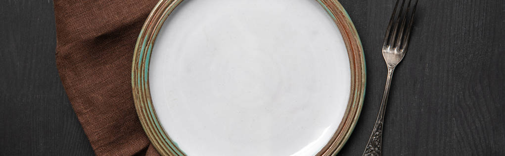 top view of vintage white empty round plate on black wooden table near brown napkin and silver fork, panoramic shot - Photo, Image
