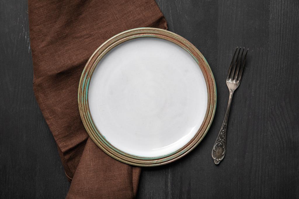 vista superior de la placa redonda vacía blanca vintage en mesa de madera negra cerca de servilleta marrón y tenedor de plata
 - Foto, Imagen