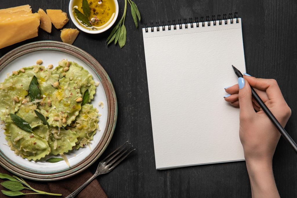 vista cortada de mulher escrevendo em caderno em branco com lápis perto servido ravioli verde
 - Foto, Imagem