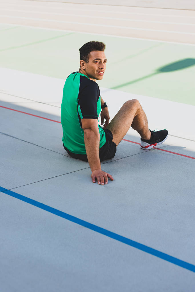 apuesto deportista de carreras mixtas sentado en pista de atletismo, sonriendo y mirando a la cámara
    - Foto, imagen