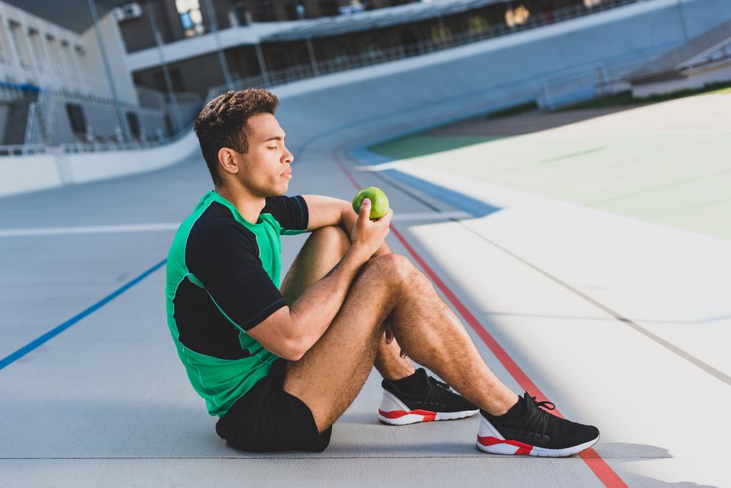 Seitenansicht des Mixed Race Sportlers, der entspannt auf der Laufstrecke sitzt und grünen Apfel betrachtet - Foto, Bild