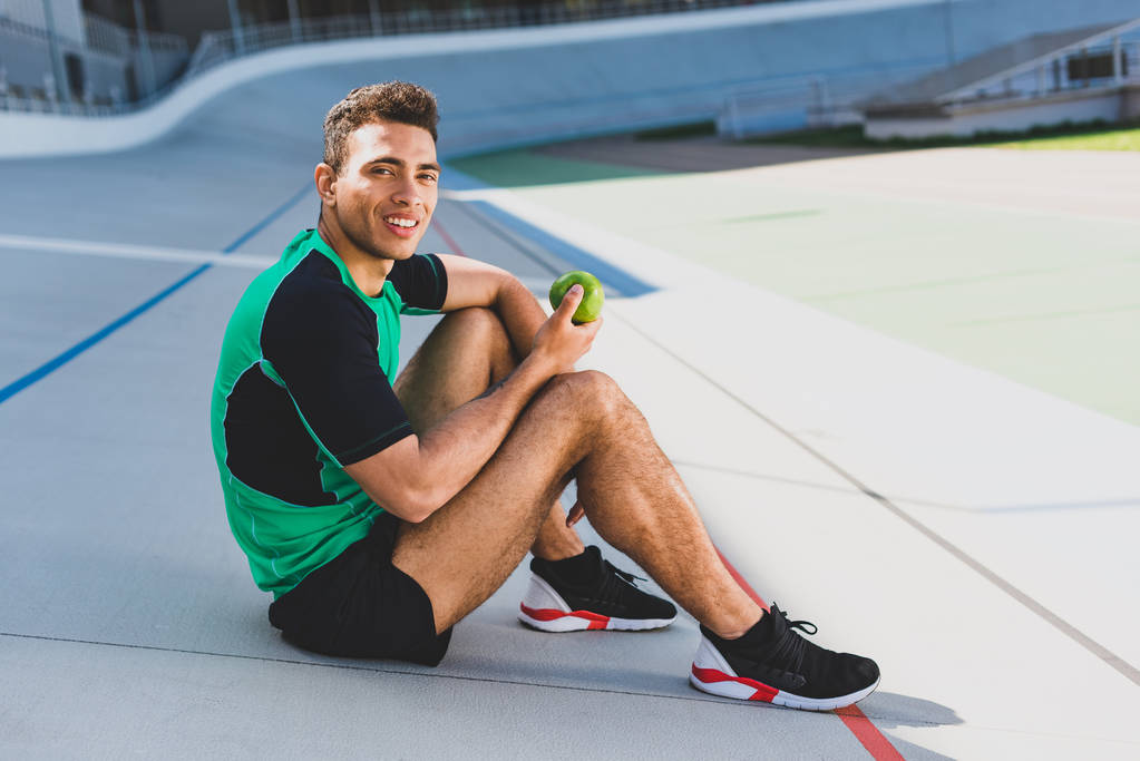 Mixed Race Sportler sitzt auf Laufbahn im Stadion, blickt in die Kamera und hält grünen Apfel in der Hand - Foto, Bild