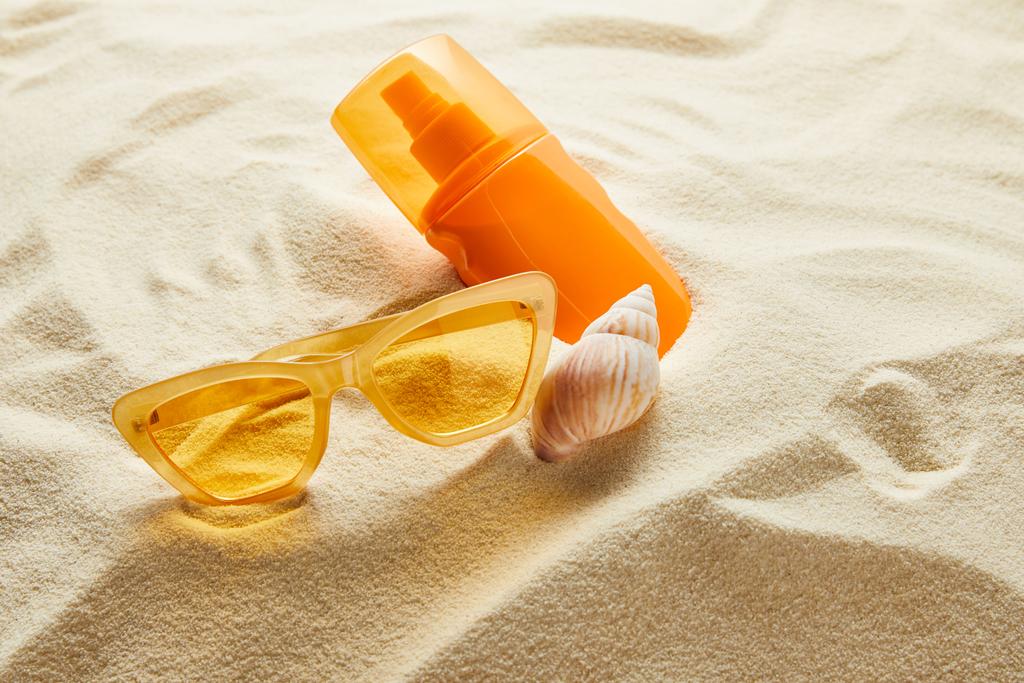 Sarı renkli güneş gözlüğü ve turuncu şişede deniz kabuğuyla birlikte kumdaki güneş kremi. - Fotoğraf, Görsel
