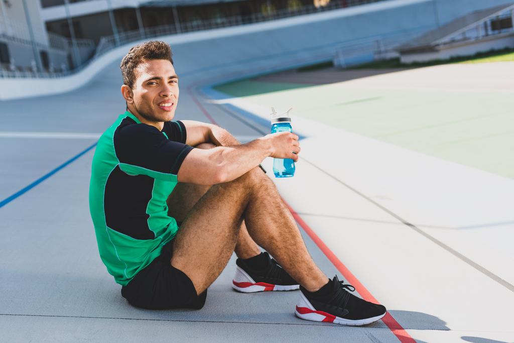 deportista de carrera mixta sentado en pista de atletismo en el estadio, mirando a la cámara y sosteniendo la botella con agua
 - Foto, Imagen