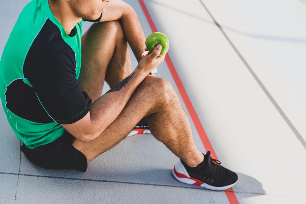 wykadrowany widok sportowca siedzi na torze i trzyma zielone jabłko - Zdjęcie, obraz